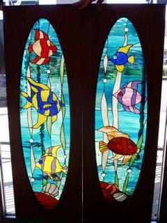 Pair of oval tropical-fish stained-glass panels in dark frames, colorful fish against aqua underwater scenes.